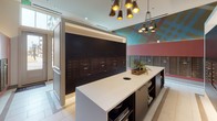 This thoughtfully designed mail room combines functionality with contemporary style. At its center sits a substantial white-topped island with built-in open shelving below, providing a convenient surface for sorting mail and storing items. A circular cutout on the countertop adds a practical detail for mail handling. The island is anchored beneath a striking multi-arm pendant chandelier with matte black and gold-interior cone shades, lending an upscale, modern aesthetic to the space.

The room's defining feature is its extensive mailbox system — banks of dark bronze individual mail compartments are mounted across two walls, set against deep charcoal panels that create a bold visual contrast against the lighter surroundings. A colorful geometric mural in teal, gold, and muted red adorns one wall, serving as a vibrant focal point that energizes the space.

Large windows on one side allow generous natural light to pour in, offering views of an outdoor terrace and neighboring architecture. The light-toned tile flooring throughout reinforces the clean, polished atmosphere. Recessed ceiling lighting supplements the natural light, ensuring the room remains bright and functional at all hours. Overall, the space balances practicality with a distinctly modern, design-forward sensibility.