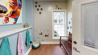 This single-floor pet care space is purpose-built for the comfort and grooming of dogs, combining functional design with a warm, welcoming atmosphere. The entry area sets a cheerful tone with whimsical paw print wall decals, playful dog-themed stickers, and a classic wood-slatted bench for seating. A white-framed window floods this area with natural light, while a wall-mounted shelf holds treats and accessories within easy reach. Teal towels hang neatly on hooks, adding a pop of color alongside a cozy pet bed resting on the hardwood-style tile flooring. The grooming area features a professional-grade stainless steel dog bathing tub equipped with a spray hose and side panels, designed for efficient and safe pet bathing. Colorful, vibrant dog portrait artwork adorns the light blue accent walls, contributing to the lively and pet-friendly ambiance. A built-in nook provides a dedicated resting or feeding station, complete with decorative murals. Grooming aprons hang near the entrance for convenience, and stainless steel water bowls are positioned on the floor. Recessed and overhead lighting ensures the space is well-lit throughout. Every detail of this space reflects a thoughtful, detail-oriented approach to pet care and grooming.