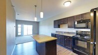 This single-floor space is designed with an open-concept layout that seamlessly connects the kitchen and living areas, creating a cohesive and functional environment. The kitchen is a standout feature, showcasing rich dark wood cabinetry that contrasts elegantly with warm granite countertops. A central island with a matching granite surface provides additional workspace and serves as a natural gathering point. Pendant lighting hangs above the island, adding both task lighting and a stylish design element. The kitchen is further equipped with black appliances, a stainless steel sink, and a classic tile backsplash that adds texture to the space. Hardwood flooring runs throughout the kitchen and living area, contributing to a warm and unified aesthetic. The living area benefits from an abundance of natural light, drawn in through a large sliding glass door that opens to an outdoor balcony or patio, offering views to the exterior. The neutral gray wall tones throughout the space create a clean, contemporary backdrop that complements the dark cabinetry and warm flooring. Overall, the space strikes a balance between modern design and everyday practicality, making it both visually appealing and highly livable.