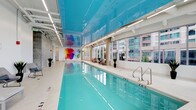 This indoor aquatic space is a standout amenity defined by its bold architectural design and vibrant aesthetic. The centerpiece is a generously sized lap pool with crystal-clear turquoise water, flanked by a clean white pool deck furnished with seating for relaxation. A glossy, vivid turquoise ceiling panel spans the length of the pool, creating a dramatic visual effect that reflects the water below and infuses the space with a sense of depth and color. On one end wall, a large-scale abstract mural bursts with multicolored splashes of red, yellow, blue, and green, serving as a striking artistic focal point. Floor-to-ceiling windows line one side of the pool, flooding the space with abundant natural light while offering views of the surrounding urban landscape. The industrial-style ceiling above features exposed elements alongside modern linear lighting, balancing raw texture with a polished finish. Potted plants are placed at intervals along the deck, introducing organic greenery that softens the contemporary environment. The overall ambiance is energizing yet sophisticated, combining functionality with thoughtful design throughout.