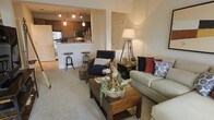 This single-floor residence showcases an open-concept layout that connects the living room and kitchen in a cohesive, functional design. The living area features plush carpeted flooring and is furnished with a neutral-toned sofa accented by navy, red, and patterned throw pillows, complemented by a dark wood armchair and a rustic wooden coffee table. A tripod-style floor lamp adds a stylish, contemporary touch to the warm ambiance.

The kitchen is positioned at the rear of the open space and features rich dark wood cabinetry, stainless steel appliances including a microwave and range, and a white breakfast bar with industrial-style bar stools, offering a casual dining option. Pendant lighting above the bar and track lighting overhead provide both task and ambient illumination throughout the kitchen area.

The neutral, light-toned walls and warm lighting create a cohesive and inviting atmosphere throughout. The open flow between the living and kitchen areas makes the space feel expansive and well-suited for both everyday living and casual entertaining.