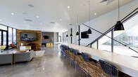 This contemporary single-floor space is thoughtfully designed to balance productivity and leisure in a sleek, open-plan environment. The interior is anchored by a long, white bar-style counter lined with modern black mesh stools, ideal for collaborative work or casual gatherings. Overhead, a row of striking black pendant lights adds a bold industrial accent, drawing the eye along the length of the counter. The polished concrete flooring throughout lends the space a refined, urban aesthetic while remaining highly durable and easy to maintain. Large floor-to-ceiling windows along one side flood the interior with natural light, enhancing the airy and expansive feel of the layout. A geometric staircase with angular black metal railings serves as a striking architectural feature near the windows. Toward the rear of the space, a lounge and recreation area is visible, complete with a wall-mounted television, brick accent wall, and a pool table, creating an inviting environment for relaxation. Recessed lighting complements the natural light throughout, ensuring the space remains well-lit at all hours. The overall ambiance is modern and energetic, combining industrial design elements with functional, multi-use areas.