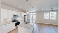 This single-floor unit offers a thoughtfully designed layout that maximizes functionality and style. The open-concept kitchen and living area serves as the heart of the space, featuring a central granite-topped island with an undermount sink and pendant lighting overhead. White shaker-style cabinetry lines the kitchen walls, complemented by a classic tile backsplash and a full suite of stainless steel appliances, including a refrigerator, range, and microwave. Light hardwood-style flooring extends throughout the main living area, adding warmth and continuity. A built-in desk nook with open shelving provides a practical workspace within the kitchen area. Large windows and a glass door in the living area invite abundant natural light and offer outdoor views. The generously sized primary bedroom features crown molding, a ceiling fan, and its own windows for natural light, with soft carpet underfoot. The walk-in closet is equipped with wire shelving systems on multiple walls, offering organized storage. The neutral color palette and clean finishes throughout create a bright, cohesive ambiance that is both modern and inviting.
