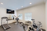 Located at 200 Pinnacle Pky in Covington, Louisiana, this fitness center offers a well-appointed workout space designed to support a variety of exercise routines. The room features crisp white walls adorned with crown molding, recessed ceiling lighting, and a durable speckled rubber flooring that provides both traction and cushioning underfoot. Natural light filters in through two windows fitted with white horizontal blinds, creating a bright and energizing atmosphere. A large wall-mounted flat-screen television is positioned above a full-width mirror, which visually expands the space and allows users to monitor their form during workouts. The gym is equipped with two treadmills, multiple Spinner-brand stationary spin bikes, a multi-function cable weight machine, and a selection of free weights including dumbbells. A wall-mounted fitness center safety rules sign is posted near the entry door, reflecting a commitment to safe usage. The single arched-panel entry door provides convenient access to the space. Compact yet thoughtfully outfitted, this fitness center delivers a functional and comfortable environment for cardiovascular and strength training activities.