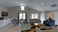 This single-floor space makes an immediate impression with its dramatic vaulted ceilings that rise to a generous peak, creating an expansive, airy atmosphere throughout the main living area. A ceiling fan is centrally mounted, complementing the recessed lighting to ensure the space is well-lit at any hour. Large windows dressed with teal drape panels line the walls, inviting abundant natural light and offering views of the surrounding exterior. French doors provide direct access to an outdoor area, further enhancing the sense of openness and connectivity to the outside. The open-concept layout seamlessly integrates a comfortable living room and a casual dining area. The living space is furnished with a neutral-toned sofa and coordinating armchairs arranged around a warm wooden coffee table, anchored by a richly patterned area rug over light tile flooring. The dining area features a classic bistro-style table with ornate chairs, adding a charming focal point. Neutral gray walls provide a clean, versatile backdrop throughout. Practical details such as a fire extinguisher and emergency exit signage are also present, suggesting the space may serve a semi-public or community function. Overall, the design balances comfort, functionality, and an inviting, welcoming ambiance.