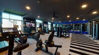 This single-level fitness center is designed with a sleek, contemporary aesthetic, characterized by dark ceilings, recessed lighting, and bold blue LED accent lighting that runs along mirrored walls. The space is organized into distinct workout zones, each thoughtfully arranged to accommodate a variety of training styles.

The main cardio area is generously proportioned, housing a full lineup of stationary bikes, treadmills, ellipticals, and stair climbers. Multiple large-screen televisions are mounted throughout, providing entertainment during workouts. Ceiling fans contribute to airflow and comfort. Large windows along the perimeter allow natural light to enter and offer views of the outdoor surroundings, creating an open, energizing atmosphere.

A dedicated strength training section features a comprehensive selection of selectorized weight machines and cable systems. Adjacent to this is a free weights area equipped with a full dumbbell rack, adjustable benches, and ample open floor space for functional movements.

A separate room serves as a functional training zone, outfitted with medicine balls, stability balls, and wall-mounted mirrors. This area features warm accent lighting and a darker, more focused ambiance.

Throughout the facility, the flooring combines textured carpet with bold black-and-white striped patterns, reinforcing the gym's modern, high-energy design identity.