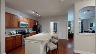 This single-floor residence offers a cohesive, modern design with rich hardwood flooring and a bold navy-and-grey color palette woven throughout. The galley-style kitchen is well-equipped with stainless steel appliances, warm cherry wood cabinetry, light countertops, and track lighting overhead. A peninsula with an integrated sink and bar seating connects the kitchen to the main living areas, creating an open, functional flow. A dedicated dining nook features a striking blue accent wall, providing an intimate setting. The living area is comfortably sized, with neutral walls and ample room for seating, while a secondary sitting area with a wall-mounted TV offers additional lounging space. Both bedrooms feature bold blue accent walls — one with carpet and a built-in desk area, the other with framed artwork above the headboard. Each bedroom is served by its own bathroom, both finished with warm cherry wood vanities, light stone-toned countertops, and large-format tile flooring. Louvered closet doors appear throughout, adding a classic, clean detail. Overall, the space presents a stylish, coordinated interior with contemporary finishes and a distinctly designed aesthetic.