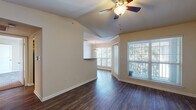 This single-story space showcases a thoughtful layout with a variety of architectural details that enhance its overall appeal. The main living area is characterized by vaulted ceilings that lend a sense of openness and volume, with a ceiling fan providing both style and practicality. Dark hardwood-style flooring runs throughout the living spaces, offering a rich, warm contrast against the neutral, beige-toned walls and crisp white baseboards.

Large windows with plantation-style blinds allow generous natural light to fill the living area, offering views of the surrounding greenery. The space opens into a defined dining nook, distinguished by a tray ceiling with crown molding and a pendant light fixture, creating an elegant transition between living and dining areas. A pass-through or countertop feature connects this zone to an adjacent area, enhancing the open-concept feel.

The bedroom features plush carpeting, a ceiling fan, and a window that brings in soft natural light, providing a comfortable and private retreat. The bathroom is efficiently designed with hardwood-style flooring, tiled walls near the shower area, and a towel bar, maintaining the cohesive neutral palette found throughout the space. Overall, the interior presents a clean, well-maintained environment with consistent finishes across all rooms.