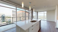Located in the heart of Portland, Oregon, this contemporary high-rise unit offers a sleek, modern living experience defined by dramatic floor-to-ceiling windows that flood the interior with natural light and frame sweeping panoramic views of the city skyline, the Willamette River, and the surrounding mountains. Rich hardwood-style flooring runs throughout, providing a warm contrast to the clean, minimalist aesthetic.

The open-concept living and dining area flows seamlessly into the kitchen, where a generously sized white quartz island with an undermount sink serves as the centerpiece. White flat-front cabinetry, a glossy subway tile backsplash, and stainless steel appliances — including a gas range, over-the-range microwave, refrigerator, and dishwasher — complete the chef-ready space. Cylindrical concrete columns add an industrial architectural accent.

The bedrooms are bright and well-proportioned, each benefiting from large windows with urban and river vistas. The bathroom features a floating white vanity with ample storage, a clean-lined mirror, and an adjoining walk-in closet. The entry foyer includes a convenient storage closet. A smart thermostat and recessed lighting are integrated throughout, lending the space a polished, move-in-ready feel.