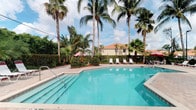 This single-story property offers a quintessential Florida living experience, highlighted by a generously sized outdoor swimming pool as a central outdoor feature. The pool, with its clear turquoise water, is framed by a spacious paved deck that provides ample room for lounging and relaxation. A set of wide entry steps leads into the shallow end, making the pool accessible and inviting.

The outdoor area is enveloped in lush tropical landscaping, with towering palm trees providing natural shade and a sense of seclusion. Colorful tropical plants and well-maintained hedgerows add layers of greenery that enhance the resort-like ambiance. Several lounge chairs and a vibrant sun umbrella are positioned poolside, offering comfortable spots for outdoor relaxation.

The building itself features a classic Florida architectural style, with a warm terracotta-toned roof and light-colored stucco exterior visible through the palms. The surrounding environment is verdant and well-maintained, reinforcing a serene, tropical setting. The open sky above, with natural sunlight filtering through the palm canopy, creates a bright and airy atmosphere throughout the outdoor space, making this property an ideal setting for those seeking a warm-weather lifestyle.
