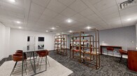 Located at 500 Merrimac Trail in Williamsburg, Virginia, this professional office suite offers a thoughtfully designed single-floor workspace that accommodates a variety of work styles and collaborative needs. The layout is comprised of several distinct areas, including an open lounge, private meeting room, and dedicated individual workstations.

The main lounge area serves as the welcoming centerpiece of the suite, featuring a high-top table with leather-upholstered bar-height chairs set atop a neutral area rug. Open-frame shelving units display books and decorative accents, while framed abstract artwork adorns the clean white walls. Recessed and drop-ceiling lighting ensures the space remains bright and functional throughout.

A compact private meeting room provides an enclosed setting for focused discussions, equipped with a round dark-topped table, green upholstered chairs, and a wall-mounted flat-screen display — ideal for presentations or video conferencing.

The individual workstation area features sleek, wall-mounted console desks in warm wood tones paired with modern bar-height chairs, set against a bold geometric-patterned accent wall in blue and grey tones. This visually distinctive backdrop adds character and energy to the workspace.

Throughout the suite, durable patterned carpet tile flooring in grey tones ties the spaces together, contributing to a polished, contemporary professional aesthetic.