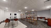 Located at 500 Merrimac Trail in Williamsburg, Virginia, this professional office suite offers a thoughtfully designed single-floor workspace that accommodates a variety of work styles and collaborative needs. The layout is comprised of several distinct areas, including an open lounge, private meeting room, and dedicated individual workstations.

The main lounge area serves as the welcoming centerpiece of the suite, featuring a high-top table with leather-upholstered bar-height chairs set atop a neutral area rug. Open-frame shelving units display books and decorative accents, while framed abstract artwork adorns the clean white walls. Recessed and drop-ceiling lighting ensures the space remains bright and functional throughout.

A compact private meeting room provides an enclosed setting for focused discussions, equipped with a round dark-topped table, green upholstered chairs, and a wall-mounted flat-screen display — ideal for presentations or video conferencing.

The individual workstation area features sleek, wall-mounted console desks in warm wood tones paired with modern bar-height chairs, set against a bold geometric-patterned accent wall in blue and grey tones. This visually distinctive backdrop adds character and energy to the workspace.

Throughout the suite, durable patterned carpet tile flooring in grey tones ties the spaces together, contributing to a polished, contemporary professional aesthetic.