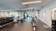 This fitness center offers a comprehensive workout facility spread across a generously sized single-floor layout. The space is divided into distinct functional zones, each designed to support a full range of exercise activities. The cardio area features an impressive vaulted ceiling with exposed wood beams and tongue-and-groove paneling, ceiling fans, and large windows that frame views of an outdoor pool and surrounding greenery, filling the space with natural light. Treadmills, an elliptical, and a stair climber are positioned to take full advantage of the scenic outlook. The strength training zone is equipped with a multi-station cable machine, adjustable benches, and a well-stocked dumbbell rack ranging across multiple weight increments. Floor-to-ceiling mirrors line one wall, visually expanding the space and providing form-checking visibility throughout. Durable rubber-tile flooring covers the entire facility, offering both safety and functionality. Overhead linear lighting ensures consistent illumination throughout. A separate utility area includes louvered closet doors, a mounted water fountain, and posted fitness center guidelines. The overall design balances practical amenity with an inviting, airy ambiance suitable for a community fitness facility.