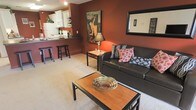 This single-floor residence features an open-concept layout that connects the living area and kitchen, creating a cohesive and functional living space. The living room is anchored by rich, deep terracotta-red walls that establish a warm, inviting ambiance throughout. Plush carpeting underfoot adds comfort, while a dark leather sofa adorned with patterned throw pillows serves as the focal point of the seating area. A wooden coffee table with a decorative woven bowl centerpiece and a side table with a classic lamp complete the living room arrangement. A large mirror and framed artwork on the walls add character and visual depth to the space.

The kitchen is visible from the living area and features white cabinetry with a dark countertop breakfast bar, complemented by bar stools for casual dining. White appliances, including a refrigerator and microwave, are neatly integrated into the cabinetry. The consistent use of warm tones throughout both areas ties the open-plan space together, creating a unified and welcoming atmosphere. Overall, the residence offers a compact yet well-appointed layout with a distinctly cozy and characterful aesthetic.
