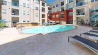 This outdoor amenity space is thoughtfully designed around a central courtyard enclosed by multi-story buildings featuring a distinctive combination of beige siding and bold red accent facades. The centerpiece is a generously sized, freeform swimming pool with clear blue water, surrounded by a stamped concrete deck furnished with lounge chairs and teal market umbrellas that provide ample shade on sunny days. Adjacent to the pool, a dedicated outdoor kitchen area sits beneath a large tensile canopy structure, equipped with stainless steel grills, dark granite countertops, and warm amber tile backsplashes, creating a fully functional space for outdoor entertaining. A secondary patio area near the building entrance features bistro-style seating with round tables and coordinating teal umbrellas, along with comfortable lounge furniture accented by teal cushions. Mature evergreen trees, blue spruce specimens in large planters, and tiered stone retaining walls filled with native shrubs and groundcover add layers of texture and natural beauty throughout the space. Flagstone and stamped concrete pathways connect each zone, enhancing both accessibility and aesthetic cohesion. The overall atmosphere is inviting, resort-inspired, and well-suited for both relaxation and social gatherings.