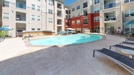 This outdoor amenity space is thoughtfully designed around a central courtyard enclosed by multi-story buildings featuring a distinctive combination of beige siding and bold red accent facades. The centerpiece is a generously sized, freeform swimming pool with clear blue water, surrounded by a stamped concrete deck furnished with lounge chairs and teal market umbrellas that provide ample shade on sunny days. Adjacent to the pool, a dedicated outdoor kitchen area sits beneath a large tensile canopy structure, equipped with stainless steel grills, dark granite countertops, and warm amber tile backsplashes, creating a fully functional space for outdoor entertaining. A secondary patio area near the building entrance features bistro-style seating with round tables and coordinating teal umbrellas, along with comfortable lounge furniture accented by teal cushions. Mature evergreen trees, blue spruce specimens in large planters, and tiered stone retaining walls filled with native shrubs and groundcover add layers of texture and natural beauty throughout the space. Flagstone and stamped concrete pathways connect each zone, enhancing both accessibility and aesthetic cohesion. The overall atmosphere is inviting, resort-inspired, and well-suited for both relaxation and social gatherings.