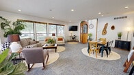 This single-floor space offers a generously sized open-plan layout that seamlessly integrates a living room and dining area into one cohesive, welcoming environment. The interior is defined by a farmhouse-inspired design aesthetic, blending rustic and contemporary elements throughout.

The living area is bathed in natural light streaming through multiple large windows adorned with wood-trimmed valances, offering views of mature trees outside. Comfortable seating, including a neutral-toned sofa accented with layered throw blankets and decorative pillows, is arranged around a wooden coffee table. Lush potted plants add vibrant greenery to the space. A wall-mounted flat-screen television is positioned for easy viewing, complemented by a decorative pine-wood cabinet nearby.

The dining area features a warm wooden table paired with a mix of metal and upholstered chairs, set atop a round area rug. Nature-inspired wall art, including an oversized landscape print and live-edge wood pieces, lends the dining zone a rustic, organic character. A decorative blanket ladder adds a charming, functional accent.

A transitional nook with floating shelves, bar-height stools, and a window provides an additional casual gathering spot. Recessed lighting throughout ensures consistent illumination, while plush carpet flooring unifies the entire space with comfort underfoot.