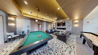 Located at 4180 Sunrise Hwy in Oakdale, NY, this well-appointed game room offers a sophisticated recreational experience within a single, generously sized floor. The space is anchored by a full-size pool table with a rich green felt surface, complemented by a sleek shuffleboard table finished in a warm wood tone. A dramatic tray ceiling with warm ambient uplighting, wood-paneled accents, and recessed fixtures creates a layered, upscale atmosphere throughout.

The lounge area features plush leather sofas arranged around a central coffee table, all set atop a bold, abstract patterned area rug that adds a contemporary flair to the space. A wall-mounted flat-screen TV is framed by an eye-catching botanical-patterned feature wall with a built-in fireplace below, providing a cozy focal point for relaxation. Sculptural pendant lighting and wall sconces in brushed gold tones add warmth and visual interest throughout.

A secondary seating nook showcases a vibrant emerald green channel-tufted sofa backed by lush indoor planters, creating a fresh, boutique-lounge vibe. A separate dining-style table with patterned chairs offers additional gathering space. Natural light filters through multiple windows, enhancing the bright, welcoming ambiance of this thoughtfully curated game room.