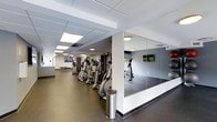 This fitness center offers a thoughtfully designed exercise space spread across a single floor. The layout is open and functional, accommodating a wide range of cardio equipment including elliptical trainers, stationary bikes, and additional strength and conditioning machines arranged in an organized row. A large floor-to-ceiling mirrored wall along one side of the room expands the sense of space and allows users to monitor their form during workouts. The flooring throughout is a durable dark rubber material, well-suited for high-traffic fitness use and providing cushioning underfoot. Overhead, a drop ceiling with recessed panel lighting ensures the space is evenly and brightly lit, creating an energizing atmosphere. Wall-mounted televisions are positioned throughout the room, offering entertainment for users during their sessions. The space also benefits from natural light through windows, contributing to an inviting and motivating environment. A dedicated open area with mirrored walls appears to serve as a stretching or functional training zone. The overall design is clean, modern, and practical, prioritizing usability and comfort for a well-rounded fitness experience.