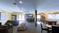 This single-floor common area is designed as a multifunctional social and recreation space. The open-concept layout is anchored by a comfortable lounge zone furnished with plush sofas and patterned carpeting, creating a relaxed setting for gathering. Recessed lighting throughout the ceiling provides consistent, even illumination across the space. A prominent wet bar or kitchenette area sits toward the center-rear, featuring a stone accent wall, wall-mounted flat-screen television, and bar-height seating with leather-style chairs — making it well-suited for casual entertaining. Natural light filters in through windows and a glass-paneled entry door, brightening the interior. To the right, a billiards table is visible, adding a recreational element to the space. Dark structural columns punctuate the floor plan without fully dividing the areas, maintaining an open and connected feel. Additional high-top seating is available near the windows, and a potted plant contributes a touch of greenery. The overall ambiance is welcoming and versatile, balancing comfort with activity-oriented amenities.