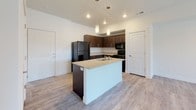 This single-floor unit is designed with a clean, contemporary aesthetic that emphasizes an open and functional layout. The living area is finished with light wood-look luxury vinyl flooring and benefits from natural light streaming through a glass-paneled front door and sidelight window, creating a bright and welcoming entry point. A ceiling-mounted light fixture and ample wall outlets add practicality to the space.

The kitchen is a standout feature, centered around a granite-topped island with a built-in sink and pendant lighting overhead. Dark espresso cabinetry contrasts with the light countertops and pale flooring, while black appliances — including a refrigerator, range, and microwave — complete the cohesive design. Recessed lighting above ensures the workspace is well-lit.

The bedroom is carpeted for comfort and includes a large sliding window that allows natural light and outdoor views. The neutral walls and ceiling-height proportions give the room a spacious, airy feel.

The bathroom is finished with wood-look vinyl flooring, a dark-cabinet vanity topped with granite, a large mirror, and a full tub-shower combination with built-in shelving. White walls and simple, modern fixtures maintain the unit's consistent, understated style throughout.