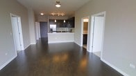 This single-floor space showcases a thoughtfully designed open-concept layout that integrates the living area and kitchen into one cohesive environment. Dark hardwood flooring runs throughout, adding warmth and a polished aesthetic that contrasts elegantly with the neutral-toned walls and white baseboards.

The kitchen is a standout feature, equipped with dark espresso cabinetry, stainless steel appliances including a refrigerator and microwave, and a light-colored breakfast bar that serves as both a functional workspace and a natural divider between the kitchen and living area. Pendant lighting above the bar adds a contemporary touch, complementing the track lighting and flush-mount ceiling fixtures that illuminate the space.

An adjacent room, accessible through a sliding or hinged door, appears to be a bathroom or utility space, fitted with matching dark cabinetry and additional lighting. The entry door, finished in a dark gray tone, provides a subtle design accent near the kitchen area.

Overall, the space presents a clean, modern aesthetic with a cohesive color palette, practical layout, and quality finishes throughout.