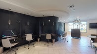 This single-floor amenity space is thoughtfully designed to serve as a multifunctional lounge and business center. The layout is anchored by large, light-toned porcelain tile flooring that runs throughout, creating a cohesive and clean aesthetic across all areas.

A standout feature is the curved, dark-walled workstation alcove, finished in deep navy or black with a dramatic wraparound desk accommodating multiple ergonomic rolling chairs. Sleek pendant lights hang at varying heights above the workstations, providing focused task lighting. A textured decorative wall panel adds visual depth to this area.

The central lounge zone features a mix of seating, including upholstered chairs and a bar-height counter with dark cabinetry and stool seating, positioned beneath a statement chandelier. A wall-mounted flat-screen television is nearby, and large windows allow natural light to filter in, offering views of the surrounding building and outdoor amenity areas.

A dedicated coffee and refreshment station is outfitted with dark wood cabinetry, dark granite countertops, a commercial-grade coffee machine, and organized storage. The entry area features floor-to-ceiling black steel-framed glass doors and partition walls, lending an industrial-modern character to the space while maintaining visual openness throughout.