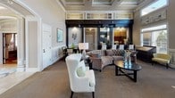 Located at 6115 Abbotts Bridge Rd in Johns Creek, Georgia, this sophisticated single-floor community lounge exudes refined elegance through its thoughtfully designed interiors. The space is defined by dramatic double-height ceilings accented with coffered detailing, lending an unmistakable sense of grandeur from the moment you enter.

A striking dark-wood fireplace serves as the room's focal centerpiece, flanked by built-in cabinetry with display shelving and decorative diamond-panel millwork that adds rich contrast to the warm neutral walls. The wainscoting that lines the lower walls further reinforces the space's classic architectural character.

Natural light floods the interior through an impressive arrangement of oversized windows and transom panes positioned high along the walls, as well as large glass-paneled entry doors that offer views of an outdoor pool area and lush greenery beyond. Multiple seating vantage points — including tufted sofas, wingback chairs, and patterned accent seating — are arranged throughout to encourage both gathering and relaxed conversation.

Neutral-toned carpeting grounds the space, complementing the dark wood accents and light walls seamlessly. An adjacent dining area is visible through the architectural passage, extending the functionality of the lounge. Overall, the space masterfully balances classic architectural detail with a welcoming, upscale ambiance.