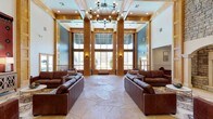 This single-floor clubhouse makes a striking impression with its grand, open-concept design. The centerpiece of the space is a dramatic great room defined by soaring ceilings framed with warm natural wood columns and beams, creating a lodge-inspired aesthetic that balances elegance with comfort. Towering floor-to-ceiling windows line the far wall, flooding the interior with abundant natural light and offering sweeping views of the surrounding landscape. The flooring features large-format natural stone tile in a neutral palette, adding texture and a sense of refined craftsmanship underfoot. A statement chandelier with exposed Edison-style bulbs hangs overhead, providing a modern industrial contrast to the rustic wood detailing and soft blue-painted walls. The lounge area is generously appointed with rich, dark brown leather sofas arranged symmetrically to encourage gathering and conversation. Recessed lighting and wall sconces complement the natural light, ensuring a warm ambiance at any hour. The overall atmosphere is welcoming and sophisticated, designed to serve as a premier social hub. Every architectural element, from the structural wood columns to the expansive glazing, works in harmony to create a space that feels both grand and inviting.