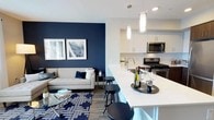 This thoughtfully designed single-floor residence combines modern aesthetics with functional living. The open-concept layout seamlessly connects the living area and kitchen, creating an inviting and cohesive space. The living room is anchored by a bold navy accent wall, wood-look flooring, and large windows alongside a glass balcony door that floods the room with natural light and provides direct outdoor access with views of the surrounding neighborhood.

The kitchen is a standout feature, offering a striking two-tone design with crisp white upper cabinetry and rich dark wood lower cabinets, complemented by white quartz countertops and a classic subway tile backsplash. A generous kitchen island with pendant lighting and bar stool seating serves as both a prep space and a casual dining area. Stainless steel appliances, including a gas range, built-in microwave, dishwasher, and refrigerator, complete the chef-ready space.

The bedroom is a calm retreat finished with plush carpeting, soft neutral tones, and a window that brings in natural light. The full bathroom features wood-toned cabinetry, a white quartz vanity, a large mirror, and a shower-tub combination, maintaining the residence's clean, contemporary style throughout.