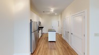 This single-floor space combines modern finishes with a clean, functional layout. The kitchen is a standout feature, outfitted with crisp white shaker-style cabinetry, dark granite countertops, and a full suite of stainless steel appliances, including a refrigerator, electric range, over-the-range microwave, and dishwasher. A movable island with a dark granite top adds additional prep and storage flexibility. Warm hardwood-style flooring runs throughout the kitchen and main living areas, complementing the neutral, light-toned walls.

The bathroom offers a polished, well-appointed design with a double-sink vanity topped with matching dark granite, large mirrors with overhead lighting, and tile flooring. A tiled shower area is also visible, maintaining a cohesive aesthetic throughout.

The bedroom features soft carpeting, a ceiling fan, and a window that allows natural light in while offering a pleasant exterior view of surrounding greenery and neighboring structures. The overall ambiance of the space is bright, clean, and contemporary, with consistent use of neutral tones and quality materials creating a cohesive and inviting environment throughout.