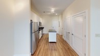 This single-floor space combines modern finishes with a clean, functional layout. The kitchen is a standout feature, outfitted with crisp white shaker-style cabinetry, dark granite countertops, and a full suite of stainless steel appliances, including a refrigerator, electric range, over-the-range microwave, and dishwasher. A movable island with a dark granite top adds additional prep and storage flexibility. Warm hardwood-style flooring runs throughout the kitchen and main living areas, complementing the neutral, light-toned walls.

The bathroom offers a polished, well-appointed design with a double-sink vanity topped with matching dark granite, large mirrors with overhead lighting, and tile flooring. A tiled shower area is also visible, maintaining a cohesive aesthetic throughout.

The bedroom features soft carpeting, a ceiling fan, and a window that allows natural light in while offering a pleasant exterior view of surrounding greenery and neighboring structures. The overall ambiance of the space is bright, clean, and contemporary, with consistent use of neutral tones and quality materials creating a cohesive and inviting environment throughout.