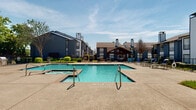 This outdoor amenity space is a standout feature of the residential community, thoughtfully designed for both relaxation and recreation. At the heart of the area lies a generously sized swimming pool with crystal-clear turquoise water, accompanied by an adjacent spa for added comfort. The pool deck is finished in a classic brick-bordered concrete surround, providing ample space for lounge seating and sunbathing under open skies.

A gated entrance with iron fencing ensures security and privacy for residents, while raised planter beds with decorative stone borders and well-maintained greenery frame the entry pathway. Mature trees and carefully curated landscaping add natural shade and visual appeal throughout the space.

A prominent wood-framed pavilion with an exposed beam ceiling and warm string lighting serves as a central gathering point, offering comfortable wicker and cushioned outdoor seating arranged for socializing. The pavilion overlooks both the pool area and a lush green lawn beyond the fencing, providing picturesque views of the surrounding buildings and landscaped grounds.

The multi-building community backdrop, featuring consistent blue-gray and brown-toned architecture with chimneys and balconies, lends a cohesive and inviting character to the overall environment. The space is bathed in abundant natural sunlight, creating a bright, open, and welcoming outdoor retreat.