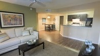 This single-story residence features an open-concept layout that seamlessly connects the living, dining, and kitchen areas into one cohesive and functional space. The living area is anchored by warm wood-style flooring and a plush area rug, providing a comfortable and inviting atmosphere. A ceiling fan overhead ensures year-round comfort, while an accent wall painted in a soft sage green adds a subtle pop of color against the neutral beige tones throughout the space.

The dining area sits just beyond the living room, highlighted by a pendant light that hangs above a bistro-style table with bar stools, creating an intimate setting for meals. Decorative touches, including wall art and a sunburst mirror, add personality to the space.

The kitchen is positioned to the right of the main living area and features dark cabinetry contrasted against light countertops and a raised breakfast bar, offering additional seating and a natural divider between the kitchen and living spaces. The overall design is cohesive and practical, with a warm, neutral palette and consistent flooring that ties each zone together seamlessly.