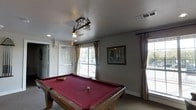 This single-floor recreational space is designed for leisure and entertainment, anchored by a full-size pool table with a rich red felt surface and solid wood frame. A decorative wrought-iron pendant light hangs directly above the table, providing focused illumination and adding a rustic, stylish touch to the room. The space is bathed in natural light, courtesy of multiple windows dressed with neutral, floor-length curtains that soften the brightness while maintaining an open and airy feel. The walls are finished in a neutral gray tone, complemented by clean white trim and baseboards that create a crisp, tidy appearance. A framed piece of wall art adds a thoughtful decorative element, contributing to the room's character. The flooring is a neutral carpet, adding comfort underfoot throughout the space. Ventilation is provided via ceiling-mounted HVAC vents, ensuring year-round comfort. An exterior door with a large window panel allows for additional light and easy access. Overall, this recreational room offers a welcoming and well-appointed environment, ideal for social gatherings and casual gameplay.