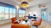 This well-appointed communal lounge and business center is thoughtfully designed to blend comfort with functionality within a single, open floor plan. The space is anchored by a striking oversized dome pendant light with a gold interior that casts a warm glow over a central seating arrangement of vibrant terracotta and blush upholstered armchairs grouped around a round white coffee table adorned with a floral centerpiece. A patterned area rug in soft gold and cream tones adds warmth underfoot, complementing the neutral carpeted flooring throughout.

Large double-hung windows dressed with blue valances line one wall, flooding the room with generous natural light and offering views of the surrounding landscape. The elevated tray ceiling, accented with recessed lighting, enhances the sense of openness and architectural refinement.

A dedicated business center zone features sleek white desks with desktop computers, task chairs, and elegant hanging pendant lights in gold and glass. An additional private workstation is positioned near the glass-paneled entry door for added convenience.

Along one wall, a well-equipped refreshment station includes light blue cabinetry, a built-in sink, a beverage dispenser, and curated wall art above — ideal for a quick break. The overall ambiance is modern, inviting, and polished, making this an ideal space for both relaxation and productivity.