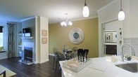 This single-story space showcases a thoughtfully designed open-concept layout that seamlessly connects the kitchen and dining areas. The dining space is anchored by rich, dark hardwood flooring that contrasts beautifully with the warm olive-green accent wall, which serves as a striking focal point. A decorative circular mirror with a layered, sculptural frame adorns the accent wall, adding an artistic touch to the room.

Overhead, a multi-arm chandelier with candelabra-style lighting provides elegant ambient illumination over the dining area, while a sleek pendant light near the kitchen zone offers additional task lighting. Crown molding runs along the ceiling perimeter, lending a refined, polished character to the space.

The dining area features a glass-top table paired with contemporary transparent chairs, offering a modern aesthetic. A white kitchen counter is visible in the foreground, topped with a decorative serving tray complete with a glass decanter and stemware. Structural columns with clean white trim define the transition between spaces, while an adjacent room — visible through an open doorway — suggests additional living areas beyond. The overall ambiance is warm, contemporary, and sophisticated.