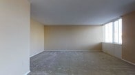 This single-floor space presents a straightforward and functional layout. The main room is generously proportioned, featuring wall-to-wall carpeting in a neutral gray tone that adds a soft, understated quality to the interior. Warm beige walls provide a clean, versatile backdrop, while smooth ceilings contribute to a sense of openness throughout. A window fitted with blinds along one wall allows natural light to filter into the room, brightening the space and creating an inviting atmosphere.

A closet off the main area offers practical storage solutions, including a full-width metal hanging rod for garments and an upper shelf for additional storage. The closet's neutral interior finish is consistent with the rest of the space, and a standard door with a brass-toned knob provides easy access. The flooring transitions to carpet at the closet threshold, maintaining a cohesive feel throughout.

Overall, the space carries a calm, neutral aesthetic that offers a blank canvas for personalization. The absence of ornate detailing keeps the focus on functionality, while the natural light and open layout contribute to a comfortable, livable environment.