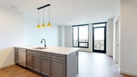 Located in Minneapolis, MN, this well-appointed one-bedroom, one-bathroom unit at 240 Portland Ave offers a thoughtfully designed floor plan with modern finishes throughout. Warm hardwood-style flooring flows consistently across the entire space, complementing the clean white walls and contemporary aesthetic.

The open-concept kitchen and living area serves as the heart of the unit. The kitchen features a large central island with a white quartz countertop and undermount sink, gray cabinetry with gold hardware, and a full suite of stainless steel appliances including a gas range, over-the-range microwave, and refrigerator. A trio of geometric gold pendant lights adds a stylish focal point above the island. Floor-to-ceiling windows and a glass door leading to a balcony flood the living area with natural light and frame sweeping views of the Minneapolis skyline.

The bedroom is a bright, comfortable retreat with large black-framed windows offering additional city views. It includes a spacious walk-in closet accessed via a sleek barn-style sliding door with built-in shelving and hanging rods.

The bathroom offers a clean, modern design with gray tile in the walk-in shower, black fixtures, and a gray vanity. A dedicated laundry room houses a stacked washer and dryer alongside built-in shelving for added storage convenience.