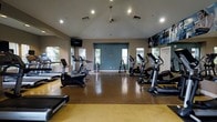 This fitness center offers a spacious, single-floor layout designed to accommodate a comprehensive workout experience. The room features vaulted ceilings that lend an open, airy feel, enhanced by a combination of recessed lighting and a central pendant light fixture. Large windows along the walls allow natural light to fill the space while providing views of the surrounding greenery outside.

The flooring consists of durable, speckled yellow and tan epoxy-style material, providing a practical and easy-to-maintain surface suited for high-traffic exercise use. Cardio equipment is the focal point, with a row of treadmills, elliptical machines, and stationary bikes arranged to maximize floor space and usability.

A wall-mounted flat-screen television is positioned for easy viewing during workouts. Full-length mirrors are incorporated along one wall, visually expanding the room and allowing users to monitor their form. A blue accent wall adds a pop of color, complementing the neutral gray and white tones throughout. Motivational fitness photography decorates the upper walls, contributing to an energetic atmosphere suited for an active lifestyle.
