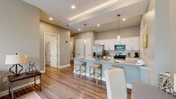 Located at 6842 Main St in Buffalo, NY, this well-appointed single-floor residence offers a thoughtfully designed layout across six rooms. The open-concept kitchen serves as the centerpiece of the home, boasting crisp white shaker cabinetry, granite countertops, and a striking two-tone island with a blue base that provides seating for multiple guests. Stainless steel appliances — including a French door refrigerator, microwave, and range — complete the chef-friendly space. Elegant pendant lighting and recessed ceiling details with tray accents add a polished touch overhead, while light hardwood flooring flows throughout the main living areas. The bright living room features a ceiling fan, sliding glass door that invites natural light, and a view of the outdoors. Two comfortable bedrooms are finished with plush carpet and generously sized windows fitted with roller shades. The bathrooms include a glass-enclosed shower with wood-tone flooring, and a separate half bath with warm neutral tones. A dedicated pantry or utility closet adds practical storage. The overall palette of soft grays and warm beiges creates a cohesive, contemporary ambiance throughout the home.