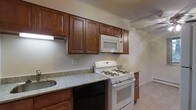 This single-floor space presents a thoughtfully arranged kitchen as its standout feature. The kitchen is outfitted with rich, dark-toned wood cabinetry that extends to the upper walls, offering generous storage capacity through multiple upper and lower cabinet configurations. The countertops display a speckled granite-style surface in neutral tones, providing durable and attractive workspace. White appliances, including a gas range, over-the-range microwave, and a black dishwasher, are seamlessly integrated into the cabinetry layout, creating a cohesive and efficient cooking environment. The sink is positioned along the counter run, adding to the kitchen's practical flow. Light-toned ceramic tile flooring runs throughout, contributing to a clean and easy-to-maintain finish. Crown molding along the ceiling adds a subtle decorative touch that elevates the overall character of the space. The neutral wall color keeps the atmosphere bright and open, allowing the warmth of the wood cabinetry to serve as the room's focal point. Overall, the space offers a functional and well-equipped environment suited to everyday living.