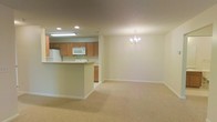 This single-floor space offers an open-concept layout that seamlessly connects the kitchen and living/dining areas, creating a cohesive and functional environment. The kitchen is positioned at one end of the space, equipped with light wood cabinetry providing ample storage, white appliances including a refrigerator, microwave, and range, and recessed and fluorescent ceiling lighting. A peninsula counter extends outward, offering a practical workspace and a natural divider between the kitchen and the main living area.

The adjoining living and dining space is generously sized, carpeted in a neutral beige tone that contributes to the overall warmth of the interior. A chandelier hangs as a subtle focal point, adding a touch of elegance to the otherwise clean and understated aesthetic. Walls are finished in a soft, neutral palette, complementing the light-toned carpet and cabinetry throughout.

The layout is well-suited for everyday living, with a logical flow between the functional kitchen zone and the open living area. The combination of recessed lighting, a chandelier, and light wall tones keeps the space feeling bright and welcoming.