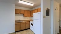 This two-floor space offers a practical and well-organized layout suited for comfortable everyday living. The kitchen is a standout functional area, featuring warm honey-toned wood cabinetry that extends along the upper and lower walls, providing ample storage space. The countertops offer generous workspace, complemented by a stainless steel sink positioned beneath the upper cabinets. White appliances, including a top-freezer refrigerator and an electric range with an oven, are seamlessly integrated into the layout, giving the kitchen a clean and cohesive look. Overhead, a large fluorescent light fixture ensures bright, even illumination throughout the cooking area. The light-toned tile flooring adds a neutral foundation that ties the kitchen together. The two-floor layout of the overall space suggests additional living areas spread across levels, offering separation between spaces for added functionality. The overall design of the kitchen reflects a straightforward, utilitarian aesthetic that prioritizes practicality and ease of use, making it a reliable and efficient space for day-to-day activities.