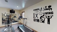 This single-level fitness room is thoughtfully designed to support a full range of exercise routines. The space features carpet flooring throughout and is illuminated by recessed ceiling lights, creating a bright and energizing environment. Natural light filters in through multiple windows with warm wood trim, adding to the welcoming atmosphere.

The room is equipped with a variety of cardio machines, including a treadmill and an elliptical trainer, both positioned for easy use and visibility of the wall-mounted television. A large wall mirror expands the sense of space and allows for form-checking during workouts. Additional equipment includes a stability ball, resistance bands, TRX suspension straps, and an assortment of kettlebells and dumbbells, making this space suitable for both cardio and strength training.

Motivational wall decals bearing words such as "Fitness," "Healthy," "Gym," and "Everyday Is a Good Day to Work Out" are displayed prominently throughout the room, reinforcing an encouraging atmosphere. A dedicated open area along one wall provides ample room for floor exercises or stretching. The room is accessed through a wood-paneled door and also includes a small waste bin and tower fan for added comfort and convenience.