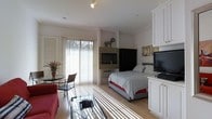 This well-appointed single-floor studio is designed to maximize both comfort and functionality within an open-plan layout. Large sliding glass doors fitted with vertical blinds flood the interior with generous natural light, casting a warm, airy atmosphere throughout the space. Light-toned hardwood flooring runs continuously across the room, visually unifying the distinct living and sleeping zones.

The sleeping area is anchored by a queen-sized bed dressed in crisp striped linens, positioned adjacent to a stone-surround fireplace that serves as a striking focal point. A sleek flat-screen television is mounted nearby, complemented by a dark armoire for additional storage. Crown molding along the ceiling lends a refined architectural detail to the space.

The living area features a comfortable red sofa and a glass coffee table, while a compact bistro-style table with wicker chairs sits beside the sliding doors — ideal for casual dining or enjoying the natural light. A television and white cabinetry unit on the opposite wall add practical media and storage functionality. Track lighting overhead provides targeted illumination, enhancing the space's versatility. Overall, the studio presents a clean, cohesive aesthetic that balances everyday living needs with a bright and inviting ambiance.