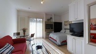 This well-appointed single-floor studio is designed to maximize both comfort and functionality within an open-plan layout. Large sliding glass doors fitted with vertical blinds flood the interior with generous natural light, casting a warm, airy atmosphere throughout the space. Light-toned hardwood flooring runs continuously across the room, visually unifying the distinct living and sleeping zones.

The sleeping area is anchored by a queen-sized bed dressed in crisp striped linens, positioned adjacent to a stone-surround fireplace that serves as a striking focal point. A sleek flat-screen television is mounted nearby, complemented by a dark armoire for additional storage. Crown molding along the ceiling lends a refined architectural detail to the space.

The living area features a comfortable red sofa and a glass coffee table, while a compact bistro-style table with wicker chairs sits beside the sliding doors — ideal for casual dining or enjoying the natural light. A television and white cabinetry unit on the opposite wall add practical media and storage functionality. Track lighting overhead provides targeted illumination, enhancing the space's versatility. Overall, the studio presents a clean, cohesive aesthetic that balances everyday living needs with a bright and inviting ambiance.