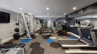 This fitness center is a comprehensively equipped exercise facility designed to support a full range of workout routines. The space is laid out across a single elongated floor plan, providing ample room for both cardio and strength-training activities without feeling cramped.

The flooring is one of the room's most distinctive design elements — a bold pattern of interlocking hexagonal carpet tiles in shades of gray, tan, and blue that adds visual energy to the space. The walls are finished in a two-tone scheme of light gray and deep navy blue, contributing to a modern, motivated atmosphere. Crown molding along the upper walls adds a refined architectural detail, while recessed lighting throughout ensures the entire space is consistently and adequately illuminated.

The cardio section houses multiple treadmills, Precor elliptical machines, and a stationary bike, positioned along the mirrored wall that visually expands the room and allows users to monitor their form. The strength-training area is well-stocked with a cable and pulley machine, a multi-functional weight rack, an adjustable bench, and a full dumbbell rack ranging across multiple weight increments. A stability ball and instructional fitness posters round out the amenities.

Natural light enters through a set of glass double doors and an interior window, enhancing the overall brightness of the space. The fitness center also includes a wall-mounted television for entertainment during workouts.