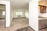 This single-floor unit offers a practical and comfortable layout designed to maximize usable living space. The living area is generously sized, featuring plush wall-to-wall carpeting and crisp white walls that create a bright, open atmosphere. A wide window allows natural light to pour in while offering views of lush greenery outside, and a wall-mounted air conditioning unit along with baseboard heating ensures year-round comfort. The living space flows seamlessly into the entryway, which transitions to durable wood-look vinyl flooring that carries through the kitchen and hallway areas. The galley-style kitchen is efficiently arranged, featuring dark cabinetry with ample storage, a countertop sink, and a full-size refrigerator, providing everything needed for everyday meal preparation. A dedicated bedroom offers generous proportions with soft carpeting underfoot and a louvered bi-fold closet door providing convenient storage. The full bathroom is cleanly appointed with wood-look vinyl flooring, a vanity sink, toilet, and a tiled tub and shower combination. Dark-trimmed doors and baseboards add a contrasting design detail throughout the unit, lending a classic and cohesive character to the overall interior aesthetic.