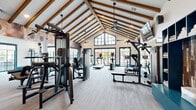 Located in Richmond, TX, this impressive fitness center offers a thoughtfully designed, single-floor space that accommodates a full range of workout options. The architectural highlight is the dramatic vaulted ceiling adorned with exposed wood beams, which lends a warm, upscale character to the facility. Large, grid-paned windows and glass doors line the perimeter, flooding the interior with natural light and offering views of the surrounding community.

The space is organized to support diverse fitness routines. A dedicated cardio zone features treadmills, elliptical machines, and upright bikes, while a strength-training area houses a comprehensive array of selectorized cable machines and multi-station units. A free weights section provides a full dumbbell rack, flat benches, and rowing machines, with a large wall mirror enhancing the sense of openness. An additional open-floor area supports functional training, equipped with cable pull systems and suspension trainers.

Throughout, light-toned textured carpet flooring provides comfort underfoot, while ceiling fans and mounted flat-screen TVs contribute to a comfortable, engaging workout environment. Teal accent walls and bold dark trim framing doors and windows add a contemporary, polished aesthetic. The facility is accessible via both double glass doors and a sliding glass panel door leading to the outdoor grounds.